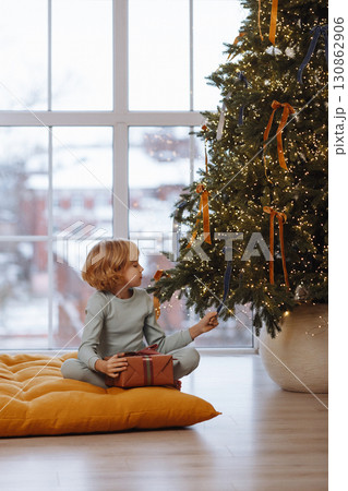 Child with gift box sitting indoors near Christmas tree lights, front view 130862906
