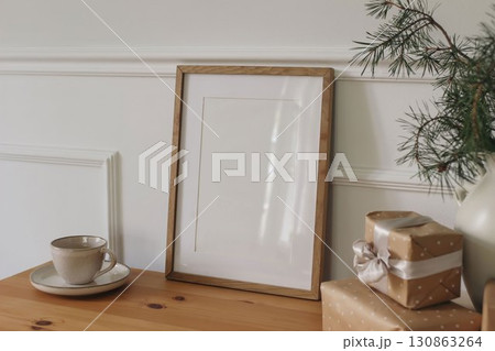 Christmas Scandinavian interior. Winter artistic composition. Blank vertical wooden picture frame mockup, cup of coffee on table. Pine tree branches in vase. Blurred gift boxes. White wall background 130863264