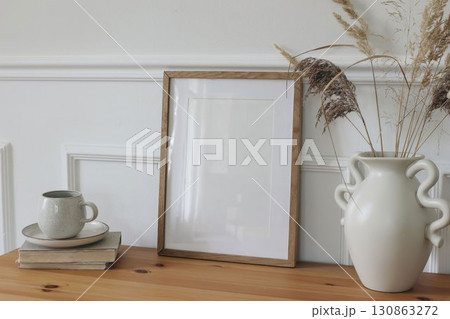 Blank vertical wooden picture frame mockup. Organic shaped vase with dry grass on table. Cup of coffee, tea on old books. Working space, home office. Art, poster display. Boho beige interior side view 130863272