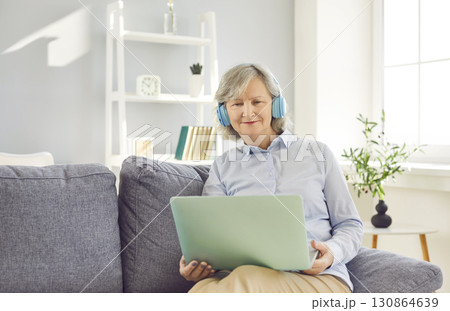 Pleasant senior woman using laptop with headphones at home siitng on sofa, smiling  130864639