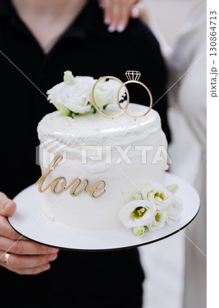 Elegant wedding cake with rings and white flowers, held by newlyweds in celebration 130864713