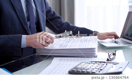 Businessman reviewing ring folder of financial documents with magnifying glass at desk in modern office. Audit and taxes in business 130864764
