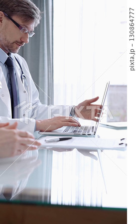 Male physician in white coat focused on laptop data in a modern clinic office while analyzing patient information on laptop. Medicine concept 130864777