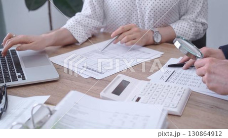Female bookkeeper or auditor is working on finances papers with a colleague, using magnificent glass, calculator and laptop. Audit and taxes Female bookkeeper or auditor is working on finances papers with a colleague, using magnificent glass, calculator and laptop. Audit and taxes 130864912