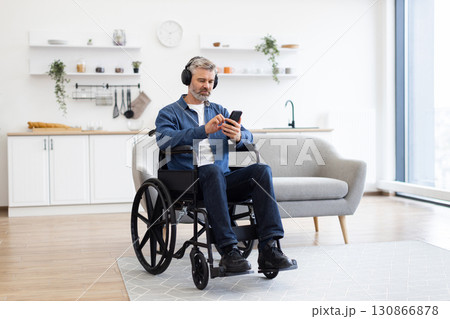 Mature Caucasian man in wheelchair enjoys music using headphones and phone in bright modern living space. Expresses passion for music, independence, and use of technology. Mature Caucasian man in wheelchair enjoys music using headphones and phone in bright modern living space. Expresses passion for music, independence, and use of technology. 130866878