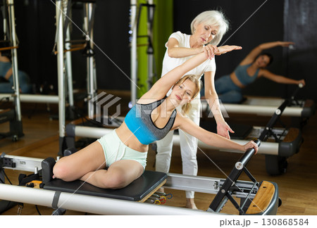 Senior woman trainer helps young woman to perform exercises in sitting position on pilates reformer 130868584