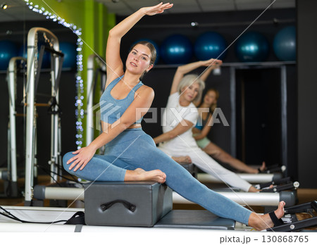 Young woman sitting on box set equipment reformer performs for abdominal muscles Young woman sitting on box set equipment reformer performs for abdominal muscles 130868765