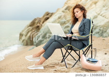 Pretty woman in office clothes, in suit, sitting on chair and working on laptop on seashore 130868837