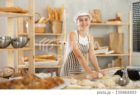Teen girl works in bakery as baker, kneads dough, works with flour. 130868893