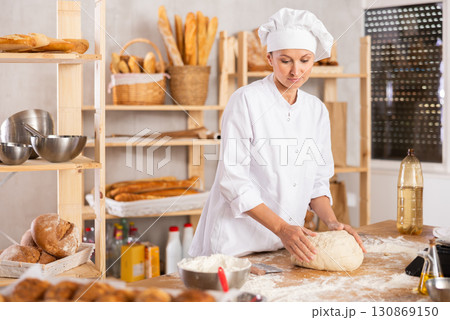 Experienced female baker preparing raw dough on kitchen table in bakery 130869150