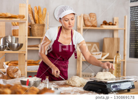 Baker woman cuts small pieces of dough from large lump and puts them on scales. 130869178
