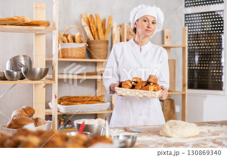 Portrait of happy female baker in white uniform working in bakehouse, holding tray with fresh bakery goods on kitchen 130869340