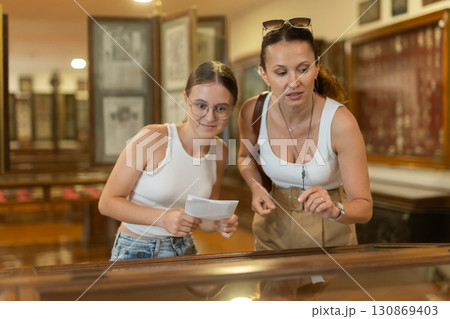 Young woman with teen daughter visitors stands near horizontal glass display case 130869403