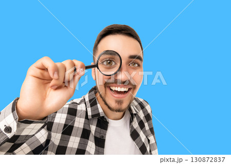 Smiling young man holds a magnifying glass expressing curiosity Smiling young man holds a magnifying glass expressing curiosity 130872837