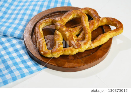 Closeup view of a pretzel loaf on a wooden tray with a blue checkered tablecloth or napkin in a white table background. Concept of Oktoberfest. Bavarian German culture festival decoration symbol 130873015
