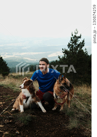 Man posing with two dogs Australian and German Shepherd in Divcibare, Serbia, with scenic mountain backdrop. Hiking with pets concept 130874259