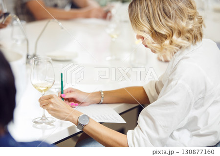 Young handsome looking woman tasting white wine and writing notes to degustation card. Professional sommelier studies wine on lecture about varieties and geographical wine. Young handsome looking woman tasting white wine and writing notes to degustation card. Professional sommelier studies wine on lecture about varieties and geographical wine. 130877160
