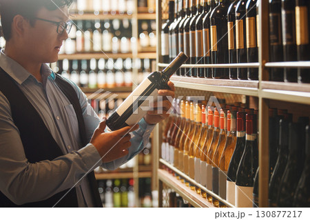 Skilled Asian Sommelier Examines Wine Bottle in Well-Stocked Cellar 130877217