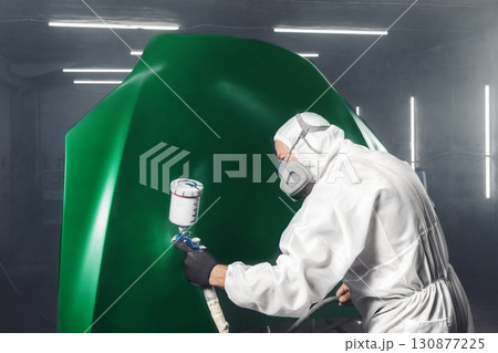 Worker paints car hood to green color in workshop chamber using airbrush. Man using spray gun during auto painting.  130877225