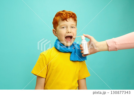 Seasonal Colds in Children Concept. Little Red Headed Boy Wrapped in a Scarf Preparing to Take Cough Throat Spray From Hands of Adult Standing on Blue Isolated Background 130877343