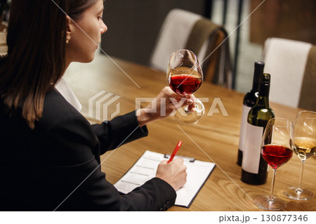 Young woman wine expert studies red wine looking on the his color sitting at the table being in cellar. Female sommelier tasting red wine during wine courses exam. 130877346