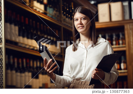 Professional caucasian woman sommelier in wine cellar making notes in tablet and choosing wine from shelve. Association of professional winemaker. 130877363