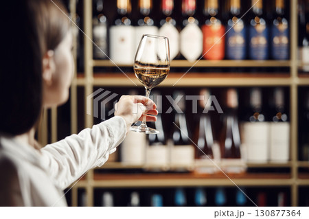 Vintner woman tasting white wine from a glass in cellar. Close up shot of female sommelier hand that hold the wine glass on shelf background. 130877364