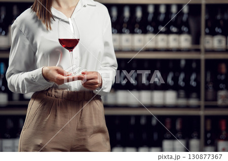 Close up shot of woman sommelier with a red wine glass stands in wine cellar with shelves full of bottles 130877367