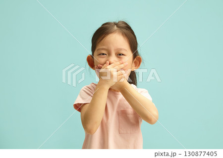 Cheerful Asian ethnicity Kid Girl with Hand Over Mouth, does not want to tell the truth, can barely contain her laughter, tries to deceive or keep silent about something standing over blue isolated. 130877405