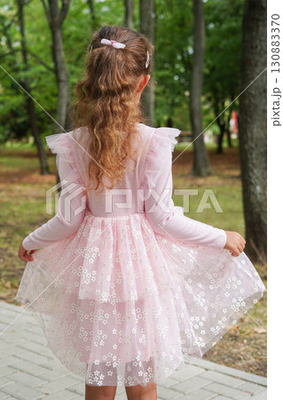 A young girl with curly hair and a bow clip is seen from behind wearing a layered pink floral dress 130883370