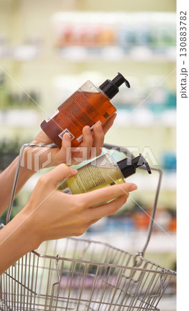 Vertical shoot. Woman selecting shampoo, body wash, hand soap in a cosmetics store, shopping basket in hand. Female customer reading labels, comparing products. Conscious choice and personal care ads 130883782