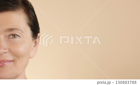 Close-up of happy female face on beige background, free space for advertising cosmetics, beauty care, skin care, spa salons. Half-face of a beautiful Caucasian middle-aged woman 50s age. Close-up of happy female face on beige background, free space for advertising cosmetics, beauty care, skin care, spa salons. Half-face of a beautiful Caucasian middle-aged woman 50s age. 130883788