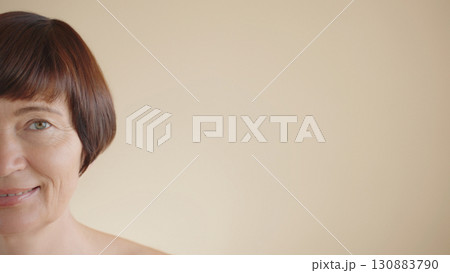 Headshot half face happy middle aged mature woman smile, brown hair beige background looking at camera copy space. Advertising concept skin care, anti-aging cosmetics products, hair care, prevention.  130883790