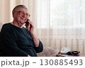 An elderly retired woman with gray hair, short haircut, wearing glasses, smiling, talking on the phone discussing health, next to a device for measuring blood pressure 130885493
