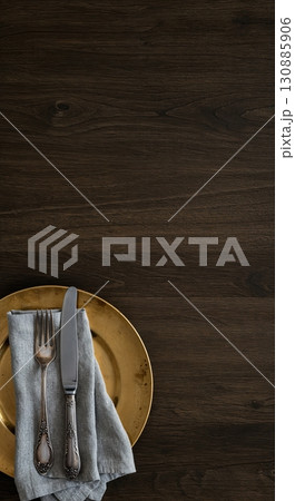 A clean, minimalist flat lay composition features a golden plate, a grey linen napkin, and ornate silver cutlery arranged on a dark rustic wooden table with ample copy space. A clean, minimalist flat lay composition features a golden plate, a grey linen napkin, and ornate silver cutlery arranged on a dark rustic wooden table with ample copy space. 130885906