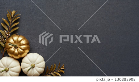 A top-down arrangement of three decorative pumpkins, one gilded gold and two pristine white, nestled with golden foliage on a dark charcoal background, offering ample copy space. 130885908