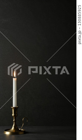 A single white candle illuminates a dark, textured background and surface, held by an elegant brass candlestick, creating a warm and contemplative atmosphere with ample copy space. 130885925