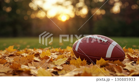 A brown American football with white laces rests on a vibrant carpet of golden autumn maple leaves with a blurred green field and warm sunset bokeh in the background. 130885933