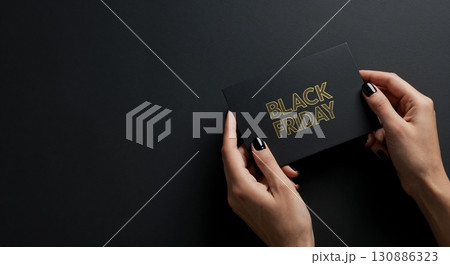 A person's elegant hands with polished dark nails carefully hold a sleek black card featuring the golden "BLACK FRIDAY" text, presented on a minimalist dark surface. 130886323