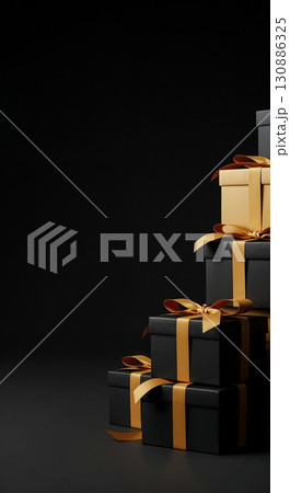 A stylish stack of black and golden gift boxes with elegant ribbons, arranged vertically on a dark surface against a deep black background, offering ample copy space. 130886325