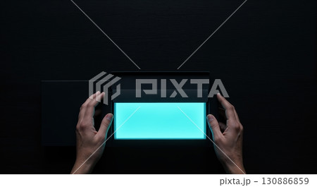 A person's hands carefully open a dark rectangular box, revealing a vibrant glowing cyan light panel from a top-down perspective on a dark background. 130886859