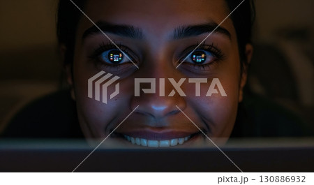A smiling young adult woman with dark hair is illuminated by the glow of a digital screen, reflecting colorful content in her wide-open eyes within a dark room. 130886932