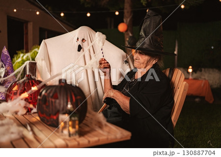 Senior woman wearing witch costume celebrating Halloween with a ghost at night 130887704
