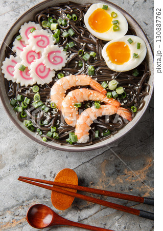 Japanese Buckwheat soba noodles with narutomaki fish roll, shrimp, eggs, sesame seeds close-up in a bowl on the table. Vertical top view 130887762