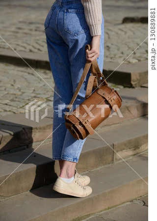 A close-up shot of a model in a knit sweater and jeans, showcasing a unique cylindrical leather bag with intricate side detailing and a belt. 130889881