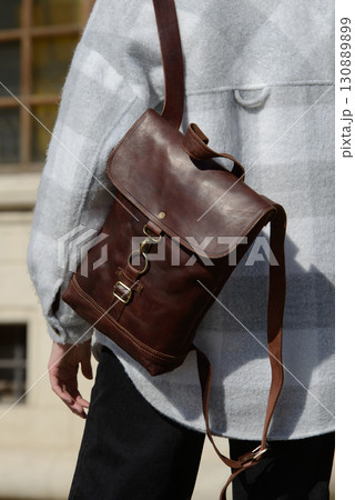 A stylish model is photographed from the back, showcasing a classic brown leather backpack and a trendy plaid overshirt against an urban backdrop 130889899
