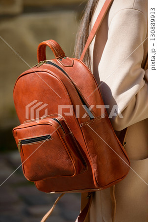 A beautiful fair-skinned model with long, dark hair is photographed from the back, showcasing a sophisticated brown leather backpack and a timeless beige trench coat. 130889913