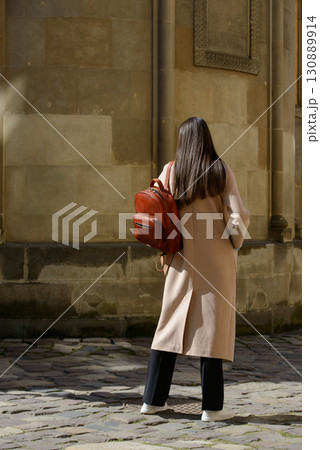 A beautiful fair-skinned model with long, dark hair is photographed from the back, showcasing a sophisticated brown leather backpack and a timeless beige trench coat. 130889914