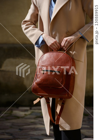 A stylish female model showcasing a brown leather backpack with detailed zippers, held against a sophisticated beige trench coat in an urban setting A stylish female model showcasing a brown leather backpack with detailed zippers, held against a sophisticated beige trench coat in an urban setting 130889915