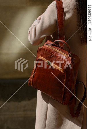 A beautiful fair-skinned model with long, dark hair is photographed from the back, showcasing a sophisticated brown leather backpack and a timeless beige trench coat. A beautiful fair-skinned model with long, dark hair is photographed from the back, showcasing a sophisticated brown leather backpack and a timeless beige trench coat. 130889931